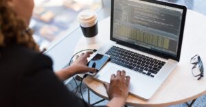 Black woman programming on a laptop with coffee, smartphone, and glasses on a desk in an office.