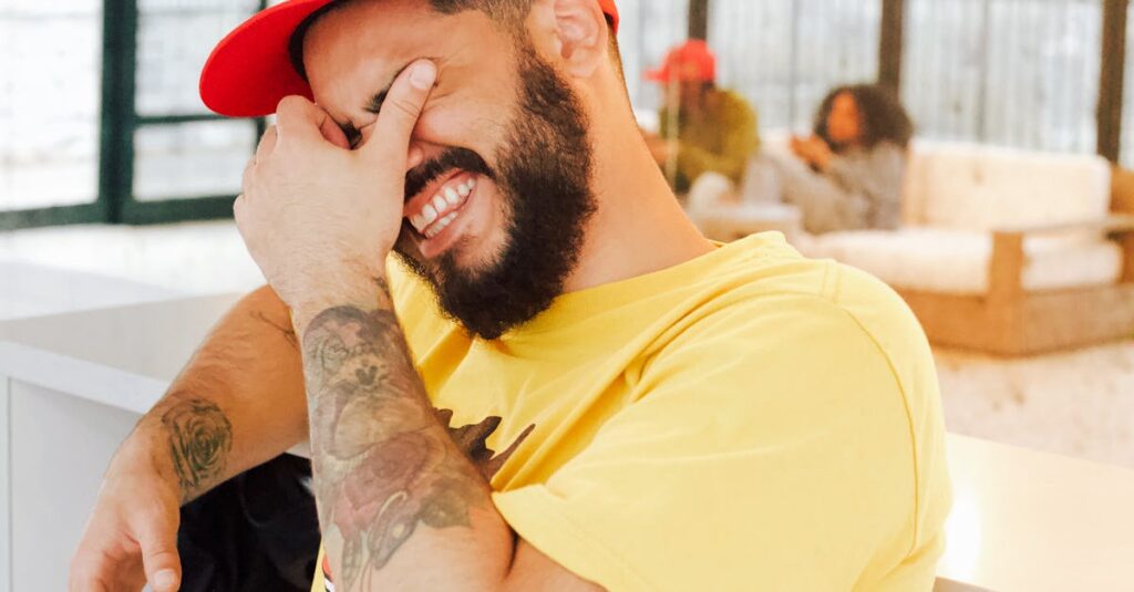 A joyful man laughing indoors with a red cap and yellow shirt, exuding happiness and relaxation.