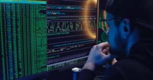 Man intently working on computer programming with code displayed on dual monitors in a dimly lit room.