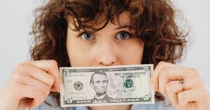 Close-up of a woman with curly hair holding a five dollar bill in front of her face.