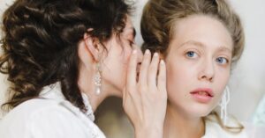 Two women dressed in 19th century fashion share a secret, showcasing elegance and friendship.